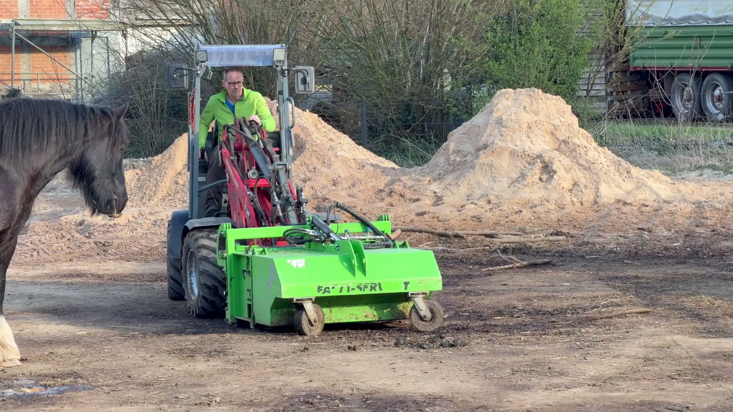 Johann Krahfuß fährt mit dem Weidemann-Hoflader und FARM-SERVANT ECO über den Sandpaddock, links ein Pferd