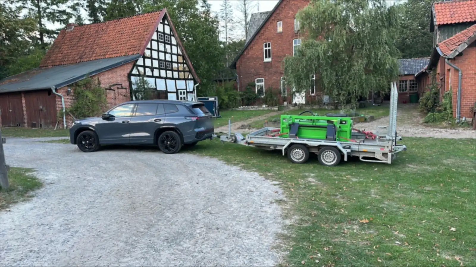 Zugfahrzeug mit Anhänger und grünem FARM-SERVANT ECO Abmistgerät auf dem Hofplatz vor einem Fachwerkhaus